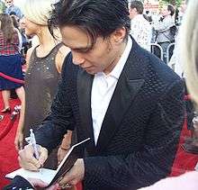 A man in a tuxedo is looking down as he signs an autograph on the red carpet.  There are people standing in the background.