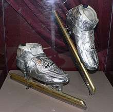 Metallic silver skates with golden blades in a glass case with the right skate being slightly elevated.  There is a burgundy curtain behind the skates.  The blades are much longer than the actual boot of the skates.