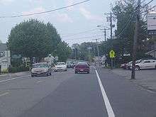 A road (NJ Route 13) packed with cars heads eastward through a residential district towards the bridge approach