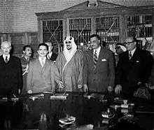 Five men standing side-by-side behind a table with documents on it. All the men are wearing suits and ties, with the exception of the man in the middle, who is wearing a traditional robe and headdress. There are three men standing behind them.