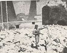 A sepia photograph shows a soldier, gun in hand, standing amidst the rubble of the destroyed synagogue. Behind him, remnants of the eastern wall show a painted fresco of Mount Sinai and two arched tablets symbolising the Ten Commandments.