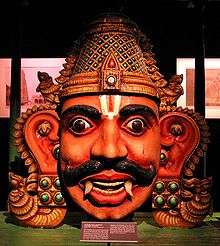 A crowned male wooden head with big eyes and ears, a Vaishnava tilak and bushy brows and large moustache. He has reddish skin, and two large canine teeth that hang down lower than his bottom lip. His eyes are wide open and he has large S-shaped ears that reach down to his chin. In the dark background, images are displayed on the back wall under lighting.