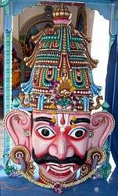 A crowned male wooden head with big eyes and ears, a Vaishnava tilak and bushy brows and moustache. A garland is seen around the neck and on the crown. He has pink skin, and two large canine teeth that hang down lower than his bottom lip. His eyes are wide open and he has large S-shaped ears.