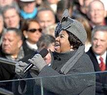 A woman her mouth open, holding a microphone. She is wearing a gray hat with a large bow, gray gloves, and a gray jacket. In the background is a crowd of people.