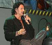 A man sitting with black shirt, black jacket, gray pants, with the left leg crossed over the right, holding a microphone in his left hand.