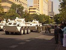 A caravan of white armoured personnel carriers rolling through a city street, with soldiers carrying body shields marching alongside. Several civilians are standing by on the sidewalk.