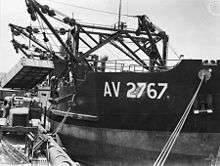 Black and white photo of a ship docked at a wharf. The ship's cranes are lifting a container above a truck.