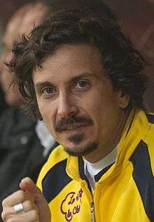 A man with long, curly hair, a moustache and a goatee in a yellow jacket