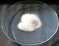 A clear glass dish on which is a small mound of a white crystalline powder.
