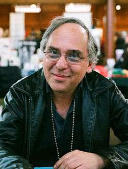 A late-middle-aged man with glasses, seated, wearing a black leather jacket, smiles at the camera.