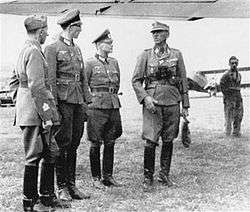 an Italian officer and three German officers in uniform standing beneath the wing of an aircraft on a grassed airfield