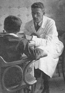 A white-coated man in his thirties sits at a table across from a boy. He looks intently at the boy through his rimless glasses. His hair is cropped fairly short on the sides and is wavy on top. The boy, seated in the foreground with his back toward the viewer, sits straight up, with one arm resting on the arm of a wooden chair.