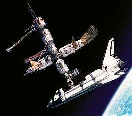 A cluster of cylindrical modules with projecting feathery solar arrays and a space shuttle docked to the lower module. In the background is the blackness of space, and in the lower right corner is Earth.