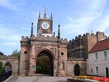 A crenellated stone arched gateway.  In the middle is a small tower with a blue clock