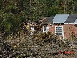 A brick home with part of its roof collapsed due to fallen trees.
