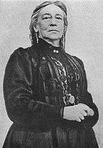 Half length portrait of a serious elderly woman in a black Victorian dress, standing with hands clasped at her waist