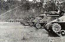 Black and white photo of a row of tanks of different designs. A line of people is visible in the background, standing in front of tall trees.