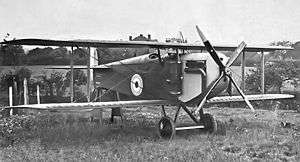 Three-quarter front view of biplane with four-bladed propeller, parked in a field