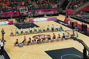 Parallel lines of two teams of players in wheelchairs, one in red, the other in green and white. They are on a basketball court, surrounded by media, official in black, and spectatotors.