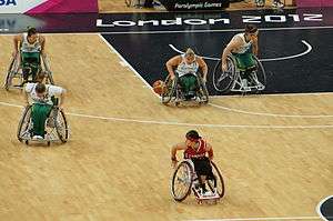 Wheelchair basketball players move across the court. Four are from Australia and one from Canada.