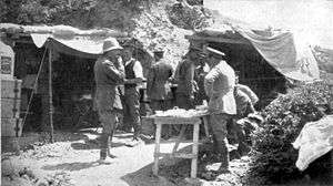 Two dugouts with tents for awnings. Wooden boxes are stacked nearby. Men in uniforms with peaked caps (and one with a solar topee) stand around a wooden folding camp table.