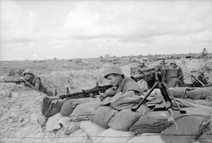Soldiers wearing helmets lie behind their weapons on a wall of sandbags, while in the background other soldiers are loading an artillery piece which is laid on a low trajectory.