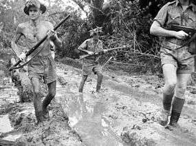 Three soldiers in shorts, wearing steel helmets but one is shirtless run along a muddy jungle track. Two carry rifles while the third has a sub-machinegun.