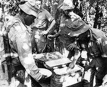 A soldier with his rifle slung is holding a plate and is being served food by another soldier while two others look on.