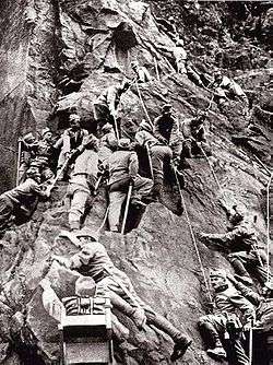 black and white photograph of men in uniforms climbing a steep rock face using ropes