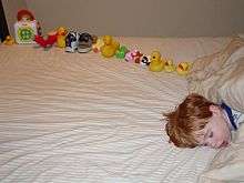 Young boy asleep on a bed, facing the camera. On the bed behind the boy's head is a dozen or so toys carefully arranged in a line.