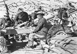 Five soldiers wearing steel helmets with a field gun. One is looking through a sight; one is loading a round; another is holding a round. The gun position is sandbagged and covered with camouflage netting.