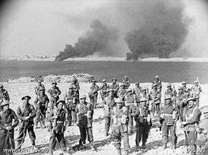 A group of soldiers stand on a foreshore. In the background, smoke billows