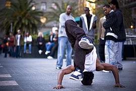 Five young men in the far background watch an African-American b-boy dance in a public plaza.