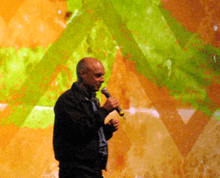 A balding man speaking into a microphone is standing in front of an abstract painting containing blotches of orange and lime green and corrugated lines.