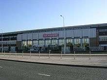 A three-storey building composed of glass windows and grey concrete stands across from a tarmac road and in front of a clear blue sky. A road passing horizontally in the foreground is divided by a pavement and metal barriers. The building's entrance appears central and is flanked by mirrored-glass windows and is marked by a red sign with the text BAE SYSTEMS.