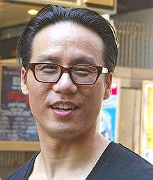 An Asian man who has short black hair and a blue shirt, and he is wearing glasses, while smiling.