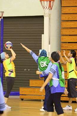 Adult women from Brunei playing netball. One team is wearing green and the other team is wearing yellow. The game is being played indoors on wooden floors. The players are all wearing pants. Some have coverings over their heads. The yellow team is in act of shooting and the green team is trying to block the shot.