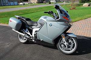 Grey motorcycle parked on an area of asphalt with red-brick paving and grass in the background