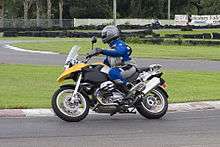  Black and yellow BMW R1200GS motorcycle, being ridden around a corner on a race track by a rider in a blue and grey suit