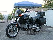 Black BMW K75T with topbox and panniers, parked on a driveway in front of a house and metal gates