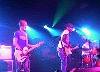 Two guitarists and a drummer are performing a song live on a stage lit by blue concert lights