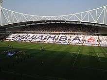 A grandstand full of people holding up coloured cards spelling out the word "Muamba" and the number 6