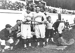 Two men smiling and holding a baseball bat, with a child next to them. Several seated men are sitting behind them, in front of grandstands.