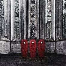 Three red coffins, closed, with kitsune (fox) masks affixed close to the top front of each. They sit below a statue with the same face as the masks, in an area that looks like a church, with stone walls and stained glass windows. The area surrounding the red coffins is in grayscale.
