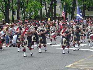 A group of men in matching outfits including kilts marches down a treelined road.