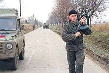 A young man wearing battledress and a beret with a Serbian flag badge stands on a road and holds an AK-47 rifle.