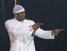 An African American man wearing white clothes and performing in a stage with a black background.