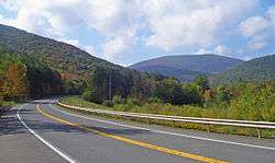 A paved highway with double-yellow centerline, shoulders and a guardrail on the far side curves across the foreground. In the distance is a high, flat-topped mountain covered in woods, with some trees showing early autumn color, framed by similar slopes on either side. Above the landscape is a blue sky with clouds.
