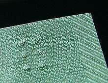 A closeup of a corner of a green banknote on a black background. To the right is the edge of the banknote, showing parallel stripes each containing various patterns (alternating printing of the numeral 20 and its textual equivalent, angled stripes, microprinted stripes). The central area depicts a patterend background of various shapes, primarily octagons and stars, forming secondary patterns such as lines, amongst which are three pairs of raised bumps.