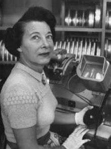 Photograph of the head and torso of a woman. She is seated in front of a Moviola machine. She is wearing white cloth gloves and is holding a reel of film. A rack with additional reels of film is visible in the background.